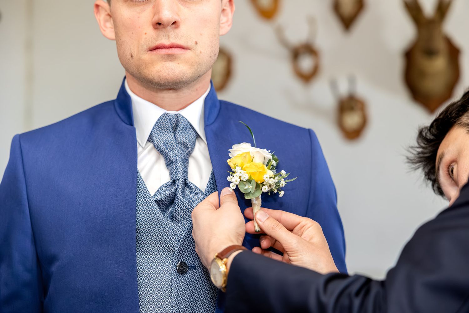 Groomsman pinning yellow boutonniere on groom's blue suit lapel.