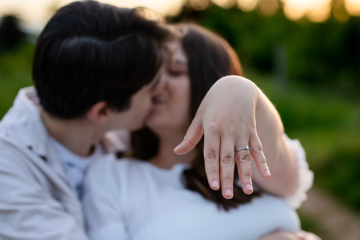 Couple kissing outdoors while woman displays engagement ring on her hand during golden hour.