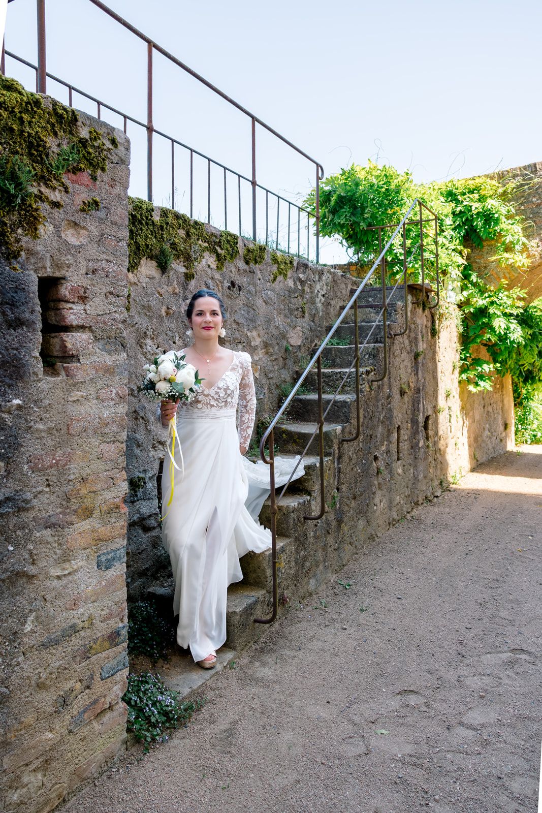 Bride in white lace gown holding bouquet poses by stone staircase at rustic outdoor wedding venue.