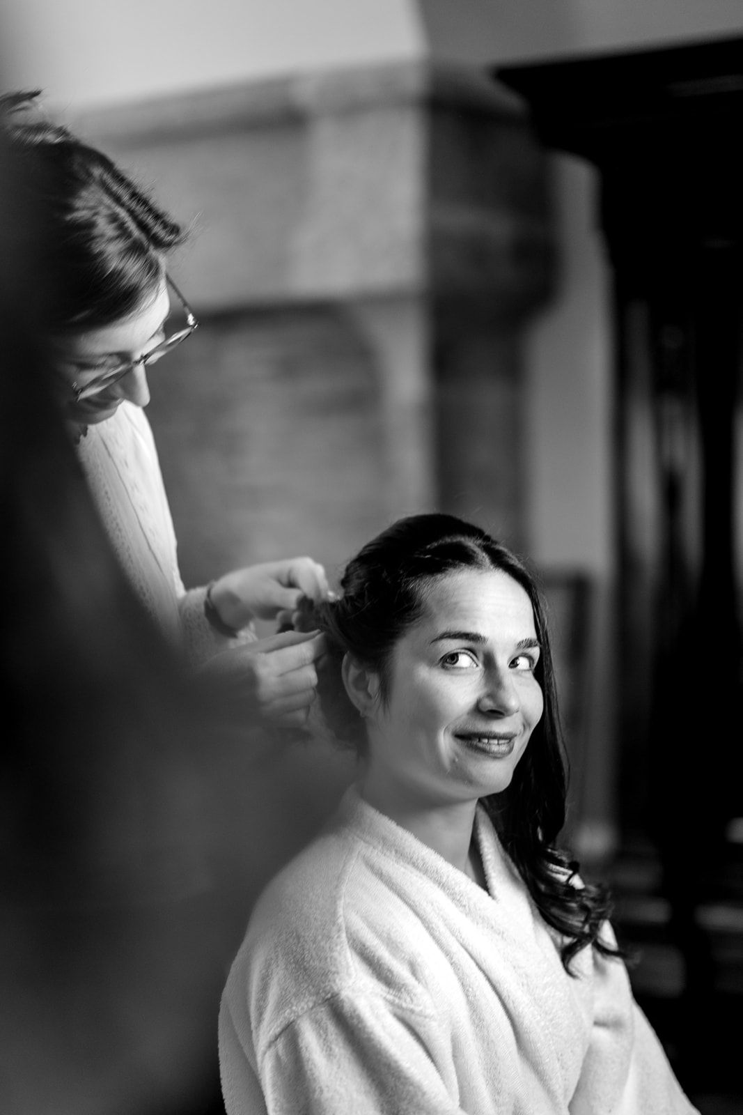 Black and white photo of a bride in a white robe smiling as a hairstylist works on her hair.