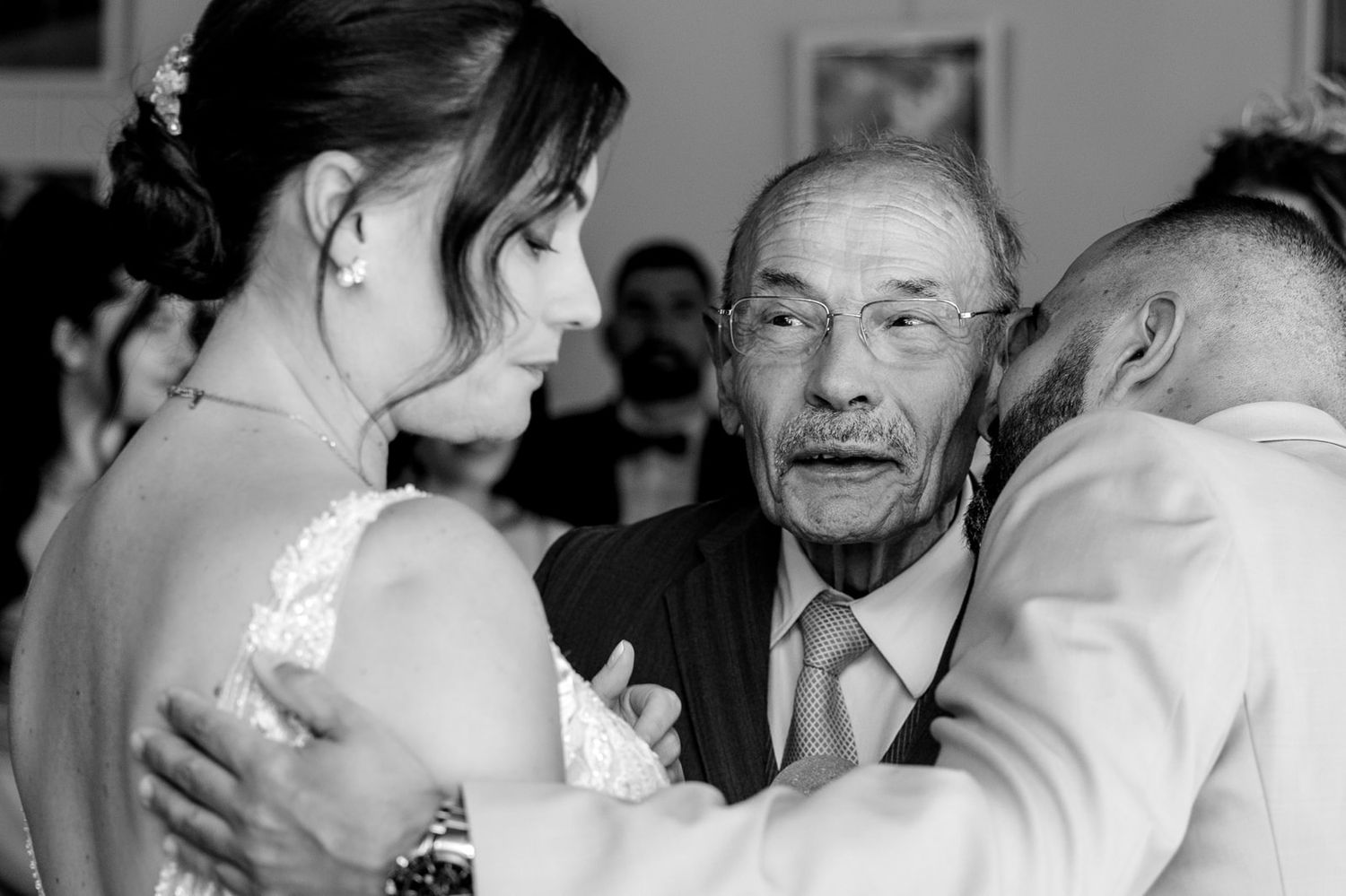 Photo de mariage en noir et blanc montrant une mariée et un homme âgé partageant un moment chargé d'émotion avec un autre invité.