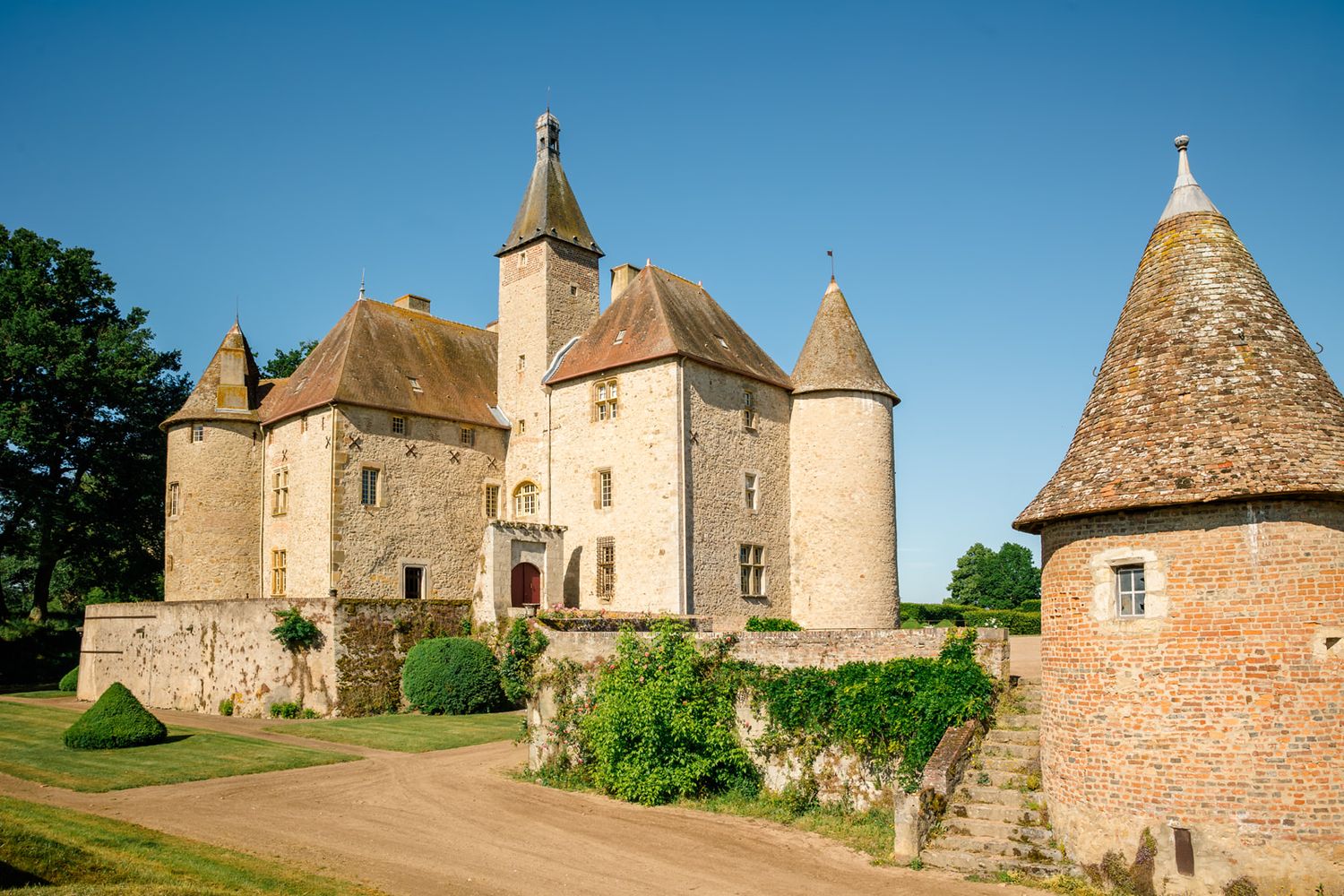 A medieval French château with conical-roofed towers, stone walls, and lush greenery under a clear blue sky.