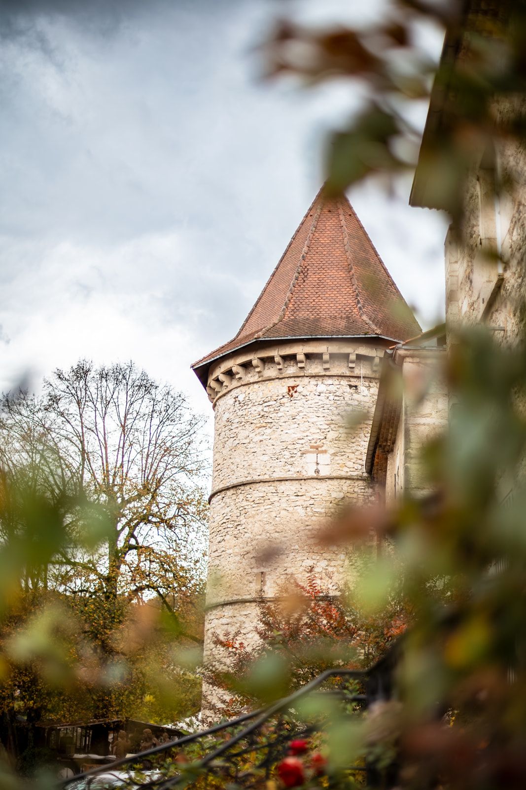 Medieval stone tower with red-tiled conical roof and circular turret rises above trees against cloudy sky.