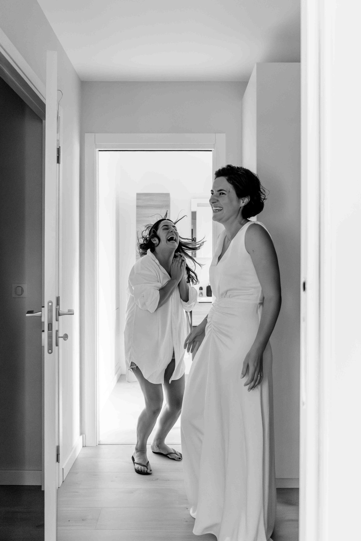A black and white candid moment showing two people in white dresses sharing a joyful moment in a hallway.