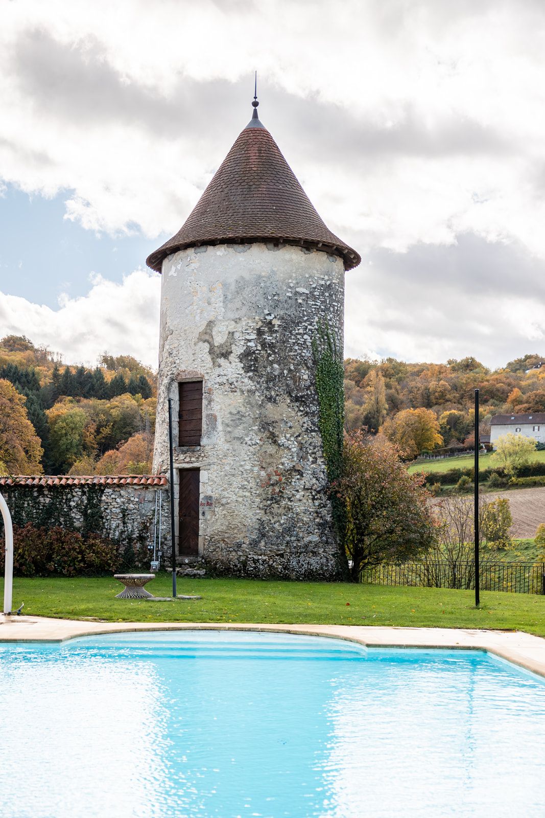 Historic stone windmill towers with conical roofs overlook a serene swimming pool in the French countryside.