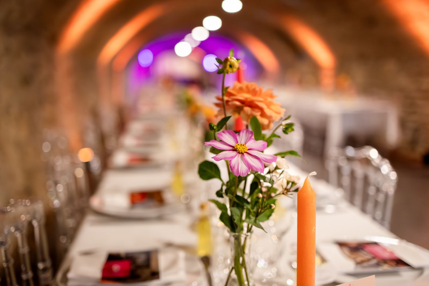 Long dining table set for a wedding reception with pink flowers in vases and warm ambient lighting.