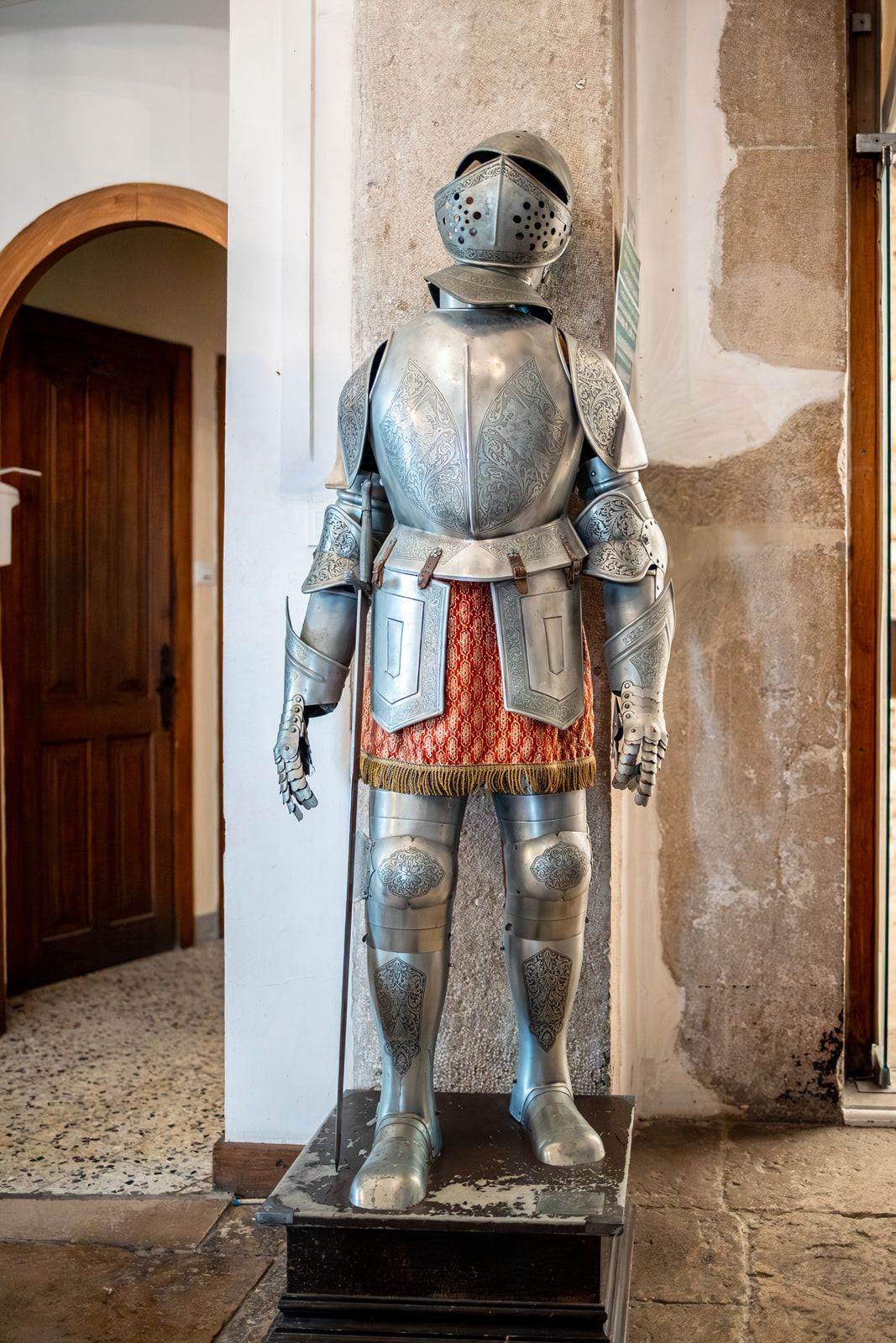 Medieval suit of armor displayed in a stone castle corridor with arched doorways and weathered walls.