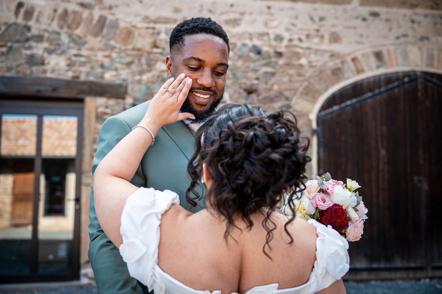 An emotional wedding moment captured outdoors with a bride holding a red and white bouquet against a rustic brick wall.