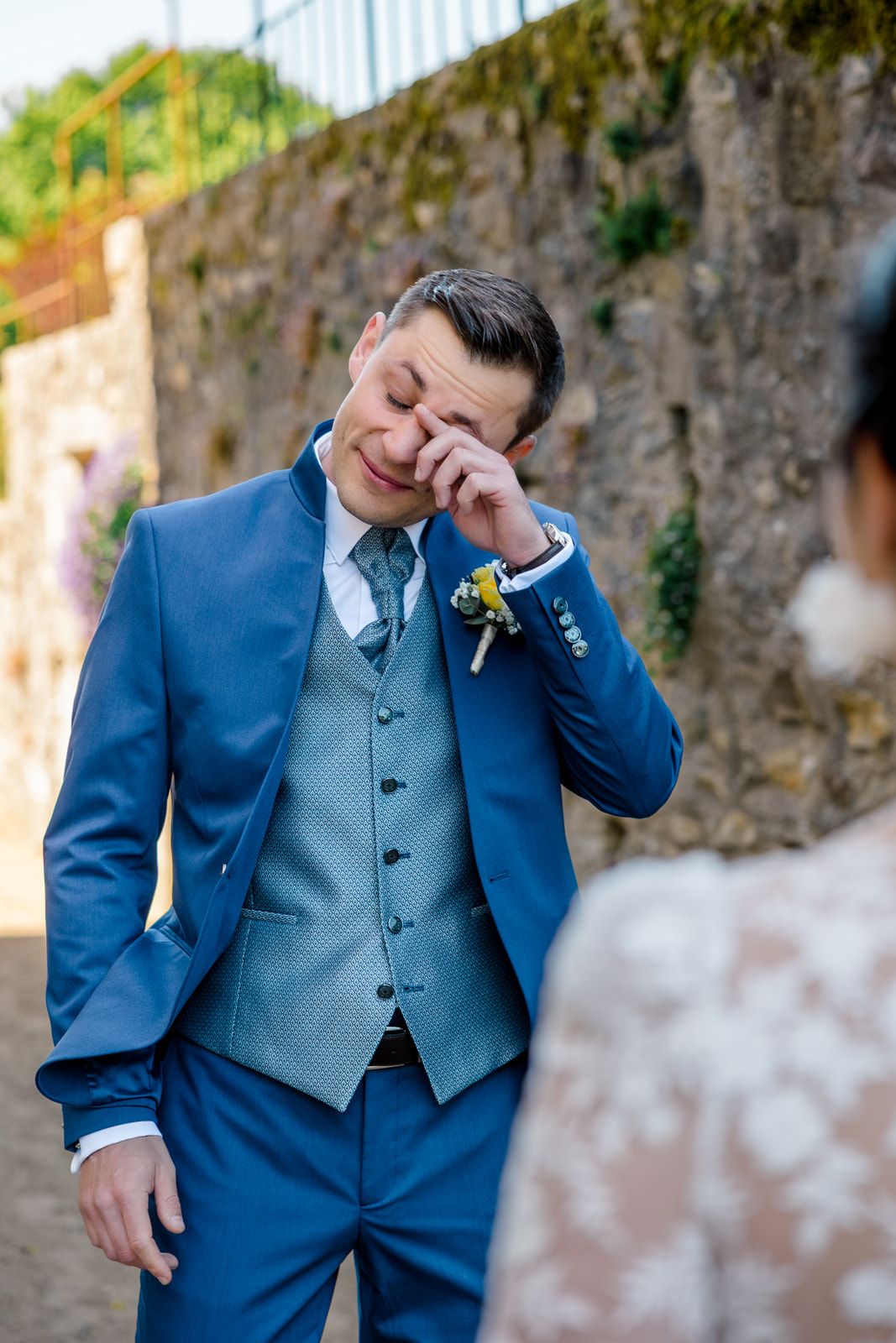 Emotional groom in blue suit wiping tears during first look at bride on wedding day.