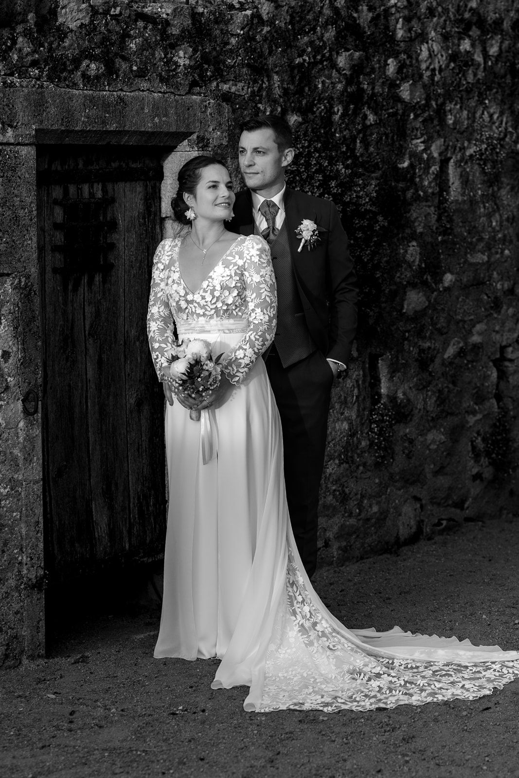 Black and white wedding portrait of bride in lace gown holding bouquet with groom in suit by rustic stone wall.