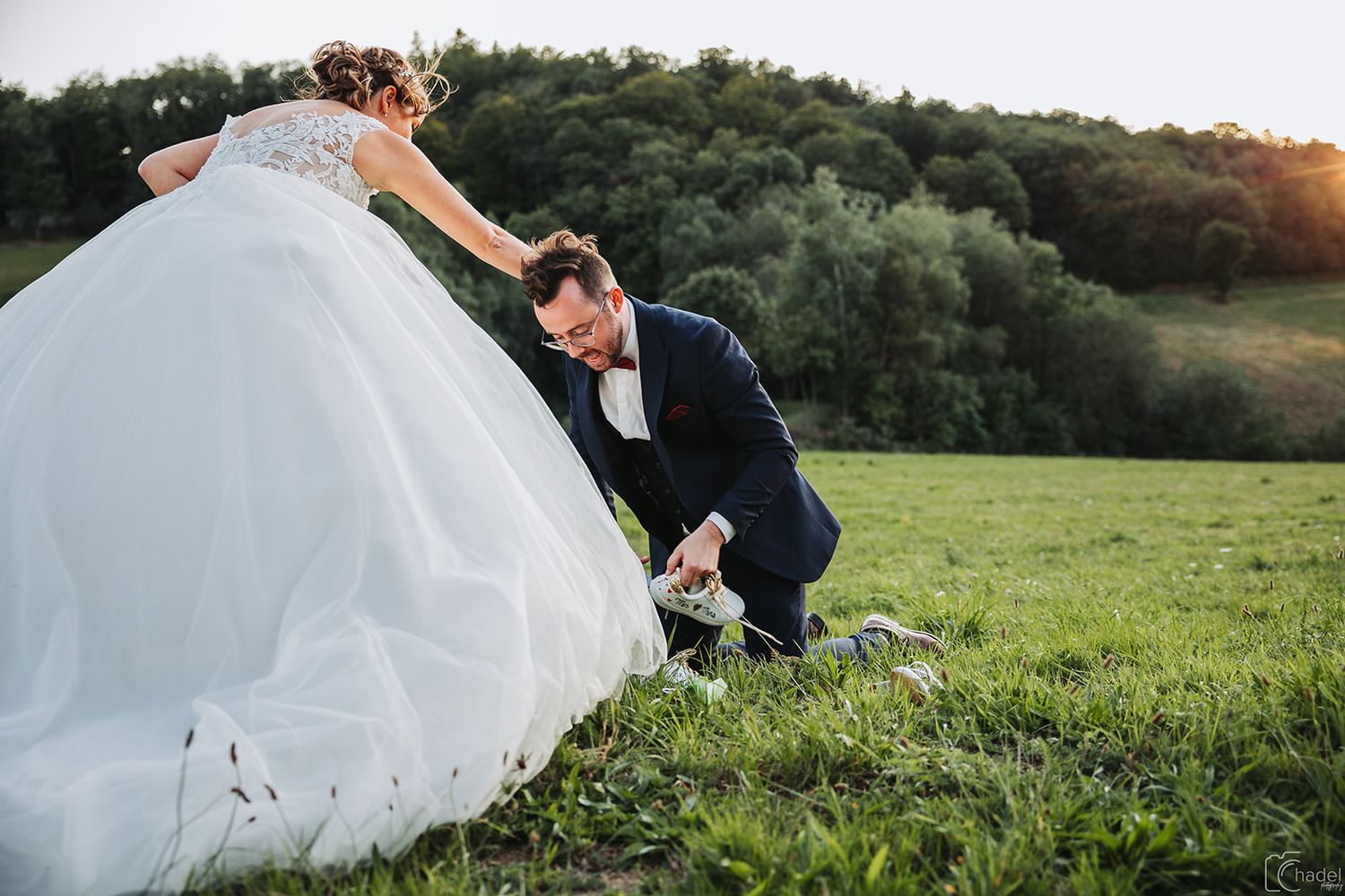 A couple in wedding attire poses together in a lush green field during golden hour sunset.