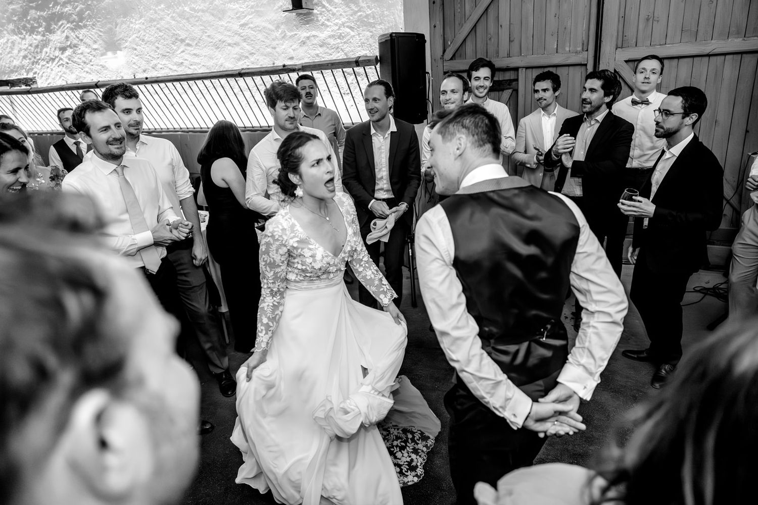 Bride and groom dance joyfully with wedding guests in black and white photo at reception.