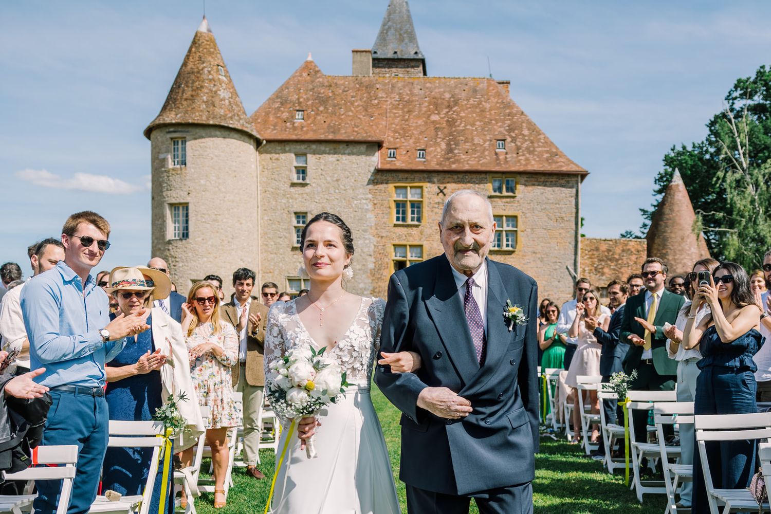 Bride walks down the aisle with elderly gentleman at outdoor castle wedding ceremony on sunny day.