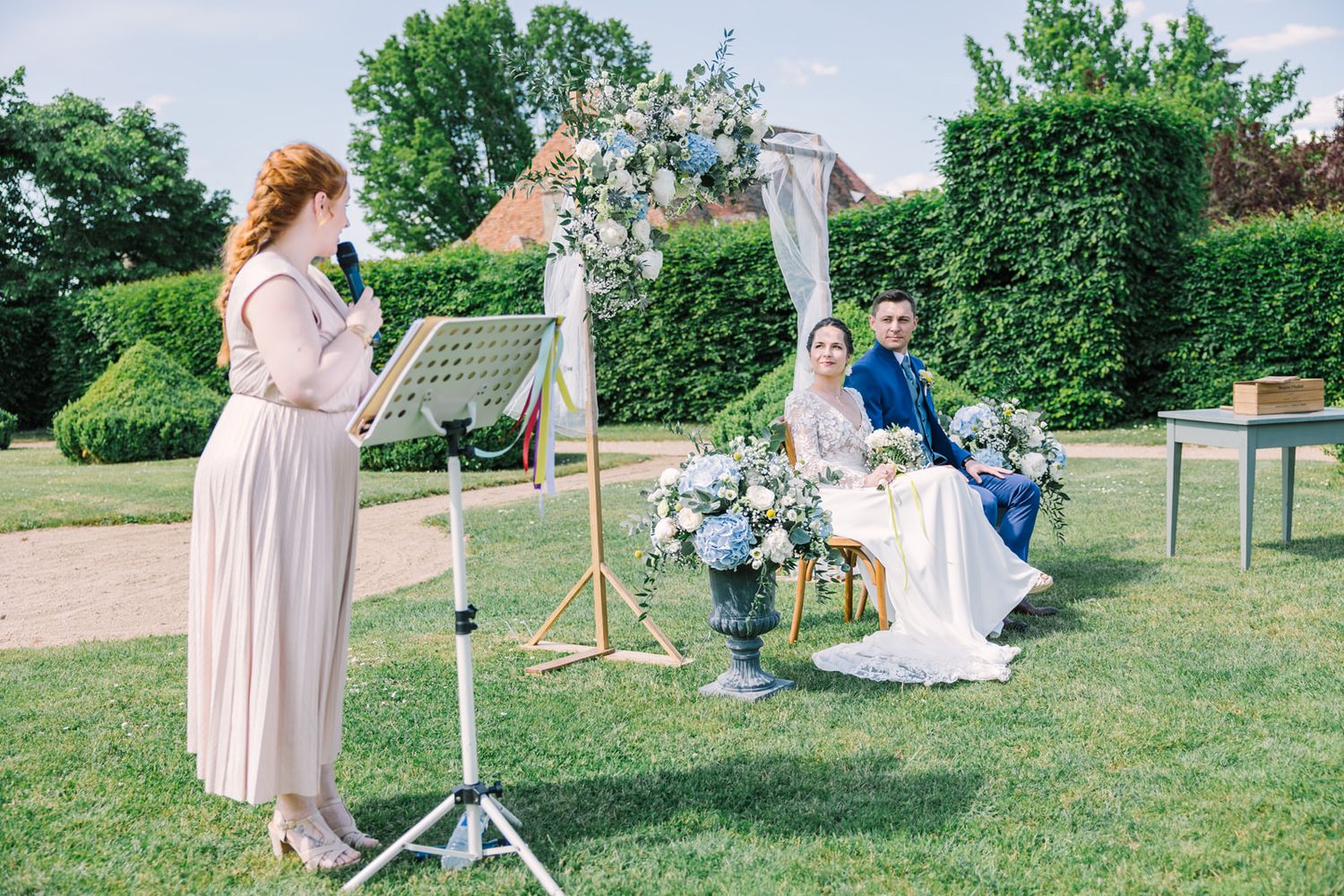 Outdoor wedding ceremony with couple seated under floral arch while red-haired officiant reads from music stand on sunny day.
