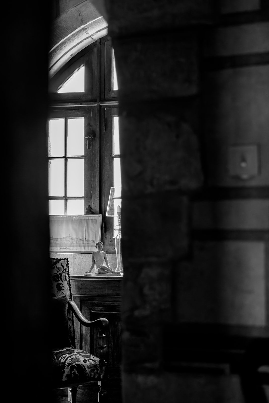 Black and white photo of a vintage room with arched window, ornate chair, and small figurine on the sill.