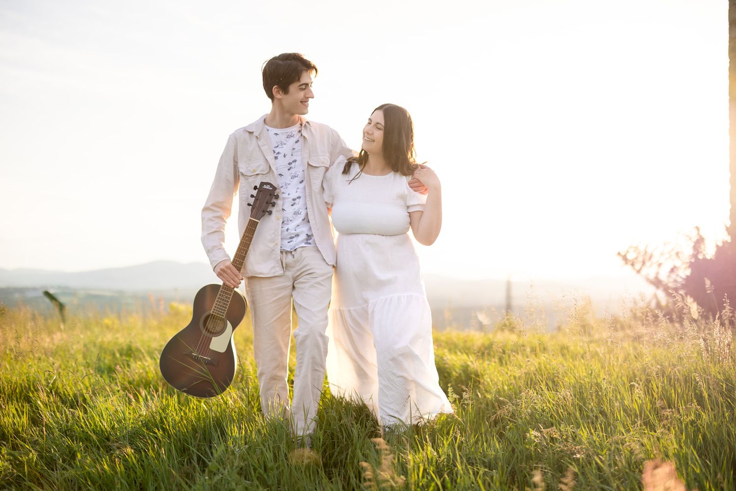 Couple walking in sunlit meadow, man holding acoustic guitar, woman in white dress, golden hour backdrop.