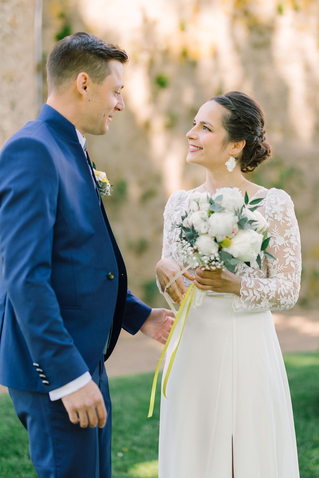 Bride in lace gown holds white peony bouquet, smiling at groom in navy suit outdoors on wedding day.
