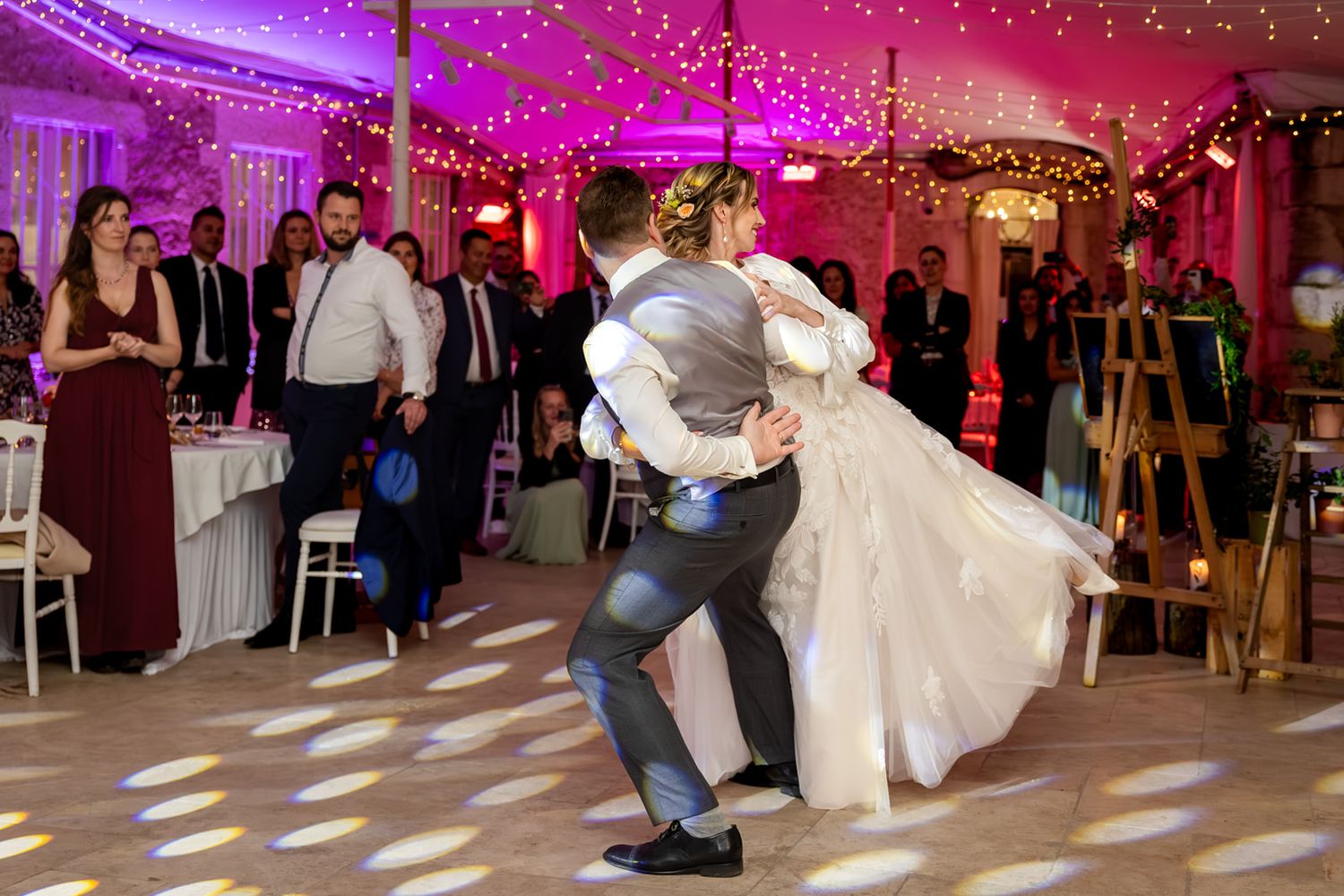 Wedding guests dance and celebrate under purple lighting and twinkling string lights at an indoor reception venue.