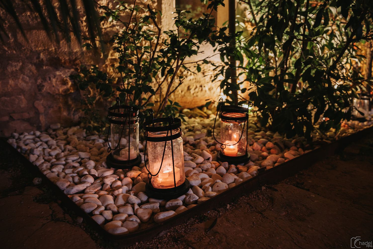 A rustic lantern with glowing candlelight illuminates a garden path lined with decorative stones at dusk.
