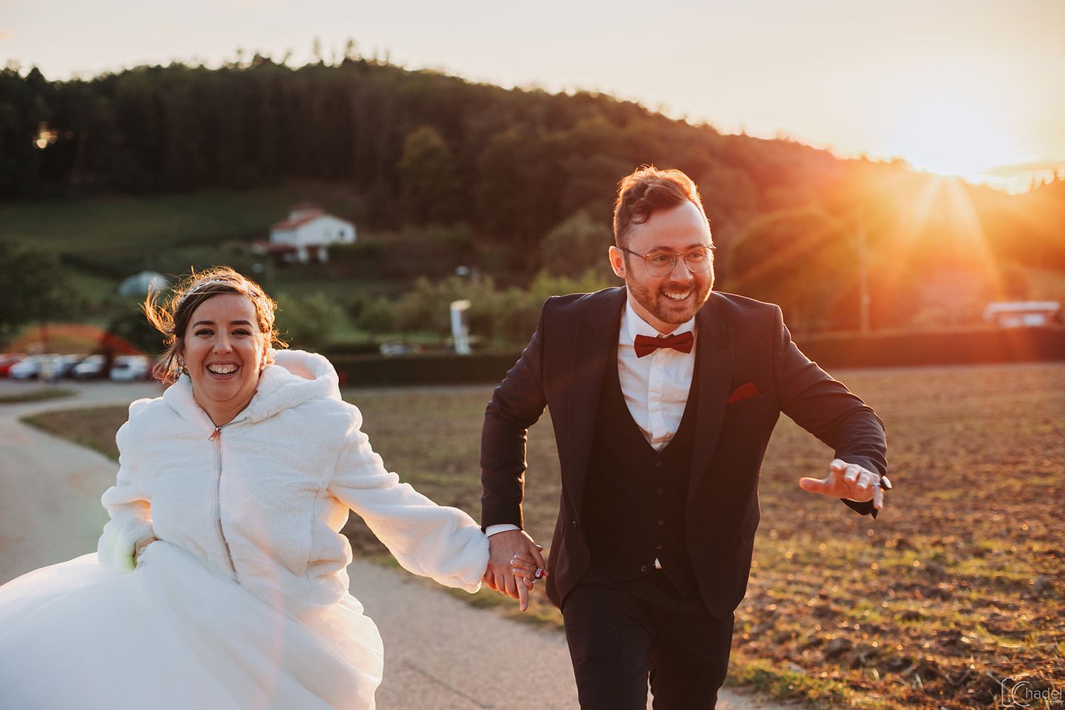 A couple walks hand in hand through a golden field at sunset while wearing formal wedding attire.