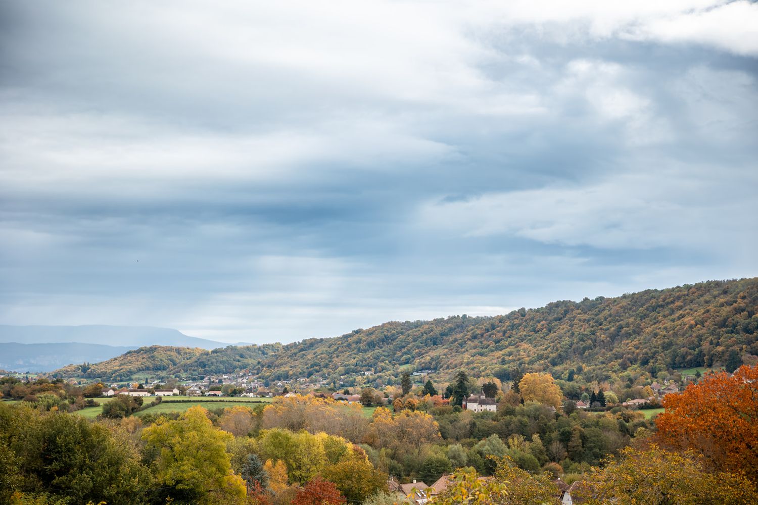 Autumn landscape view of rolling hills with fall foliage and scattered houses under cloudy mountain skies.
