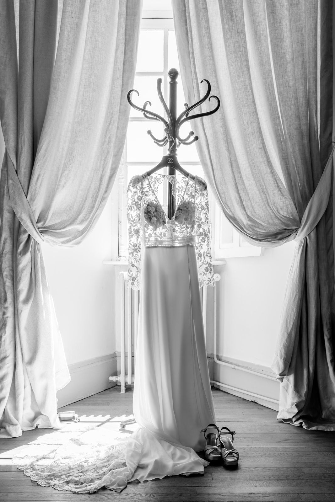 Black and white photo of an elegant wedding dress hanging on a coat rack between draped curtains with heels on the floor.