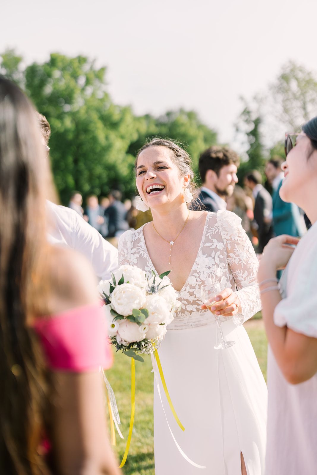 A joyful bride in a lace gown holds a white bouquet, laughing with guests at an outdoor wedding celebration.