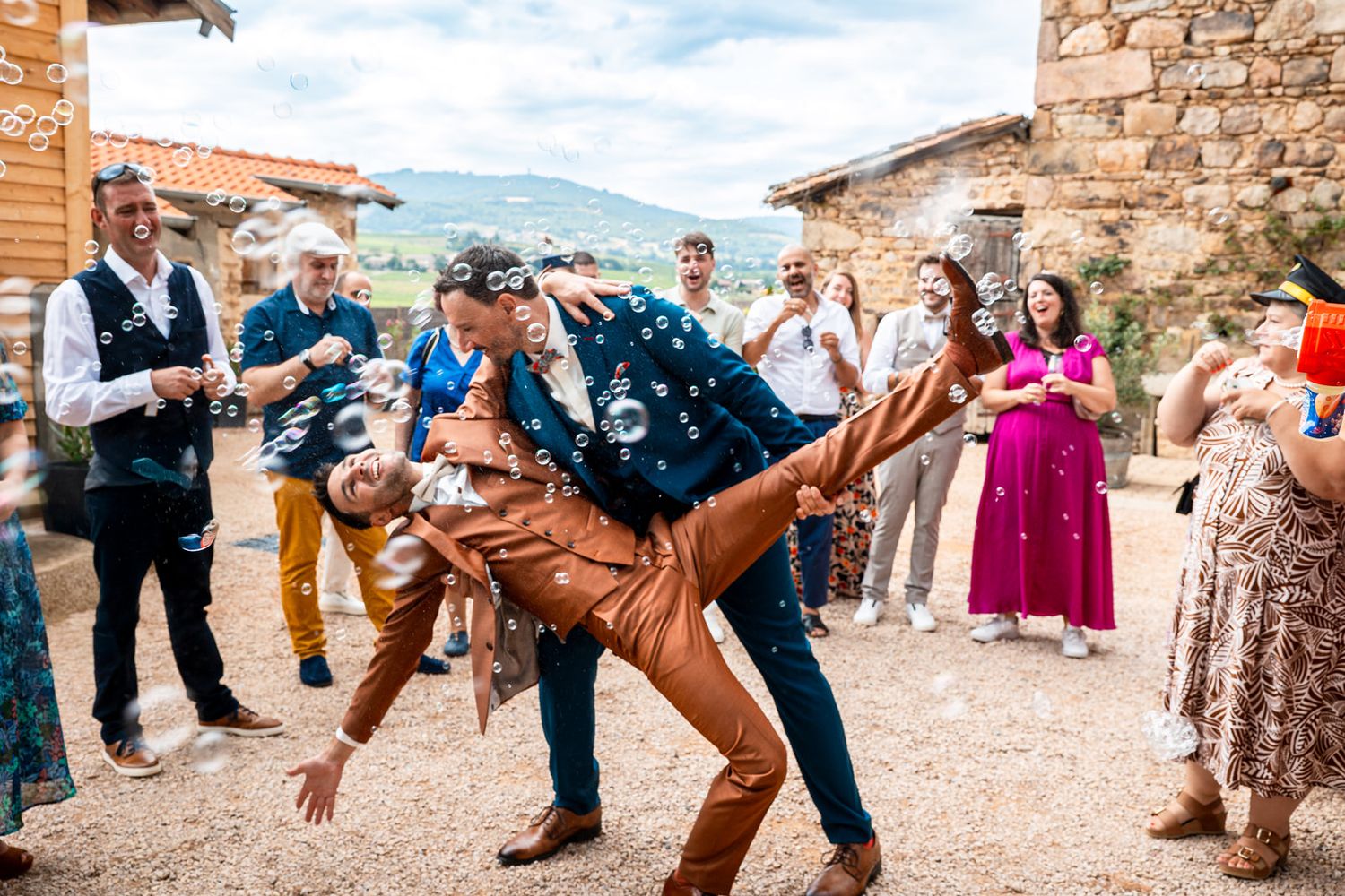 Wedding guests dance in a line at an outdoor venue with stone walls and mountain views in the background.