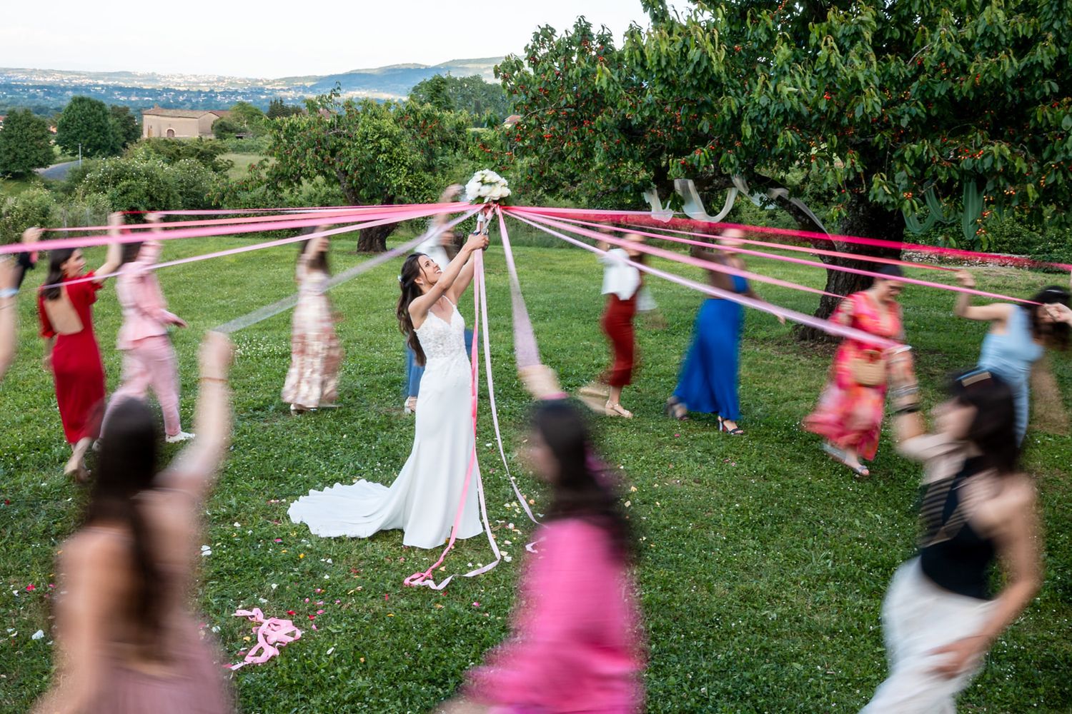 Wedding guests form a circle on a green lawn holding pink ribbons during an outdoor celebration ceremony.