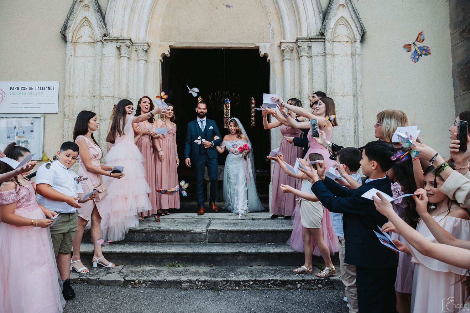 Wedding party members in pink dresses throw flower petals as newlyweds walk through outside a stone building.