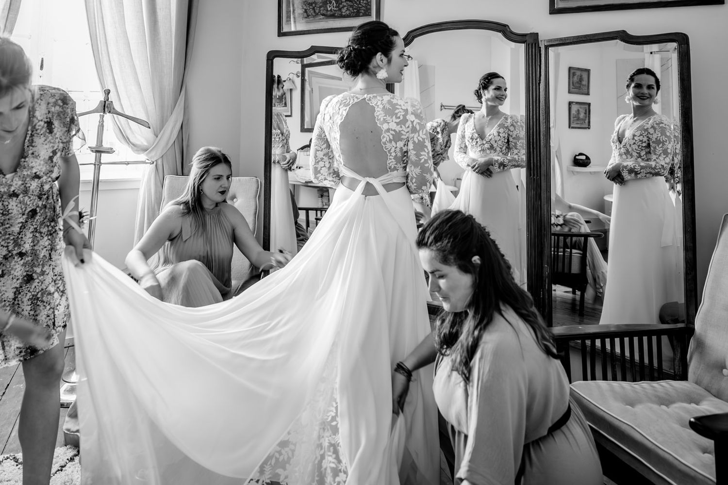 Bridesmaids helping bride into white lace wedding gown, captured in black and white with mirror reflections.