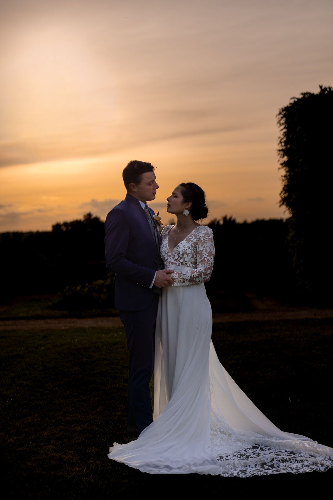 Bride and groom share a romantic moment at sunset, silhouetted against a golden orange sky in wedding attire.