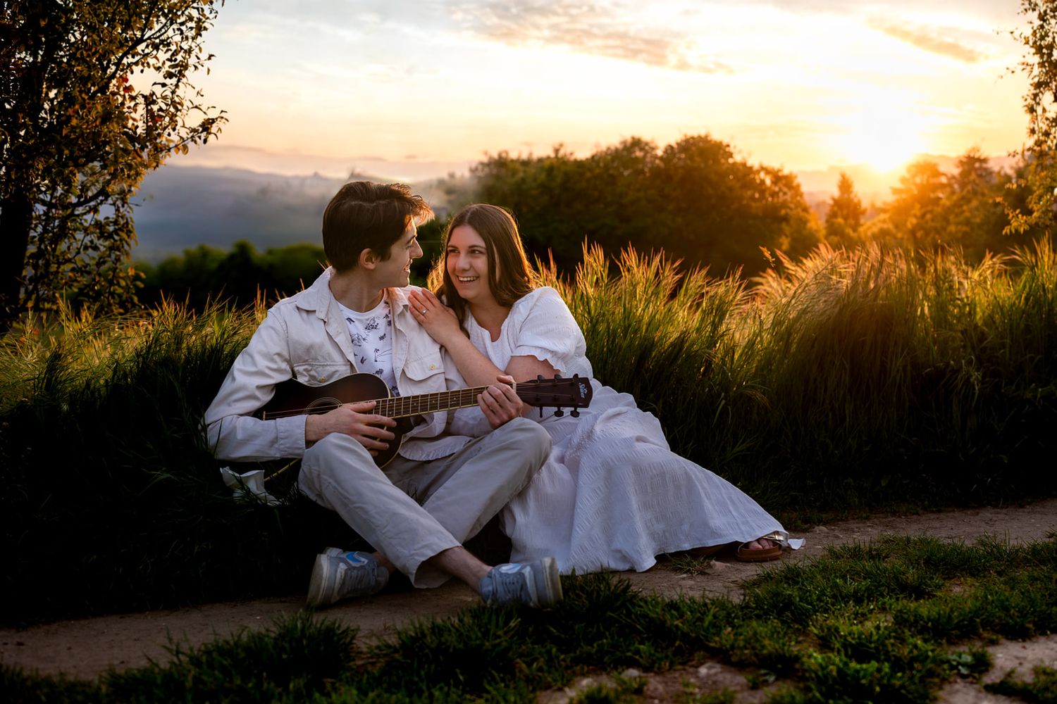 Couple sitting outdoors at sunset, man playing ukulele, woman in white dress smiling, golden hour light.