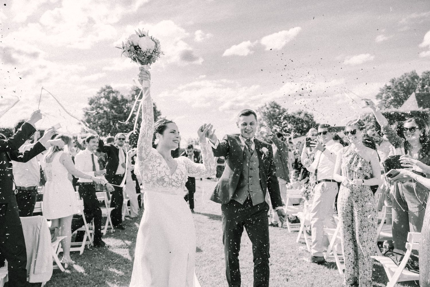 Bride and groom walk through confetti shower after outdoor wedding ceremony, guests cheering in black and white photo.