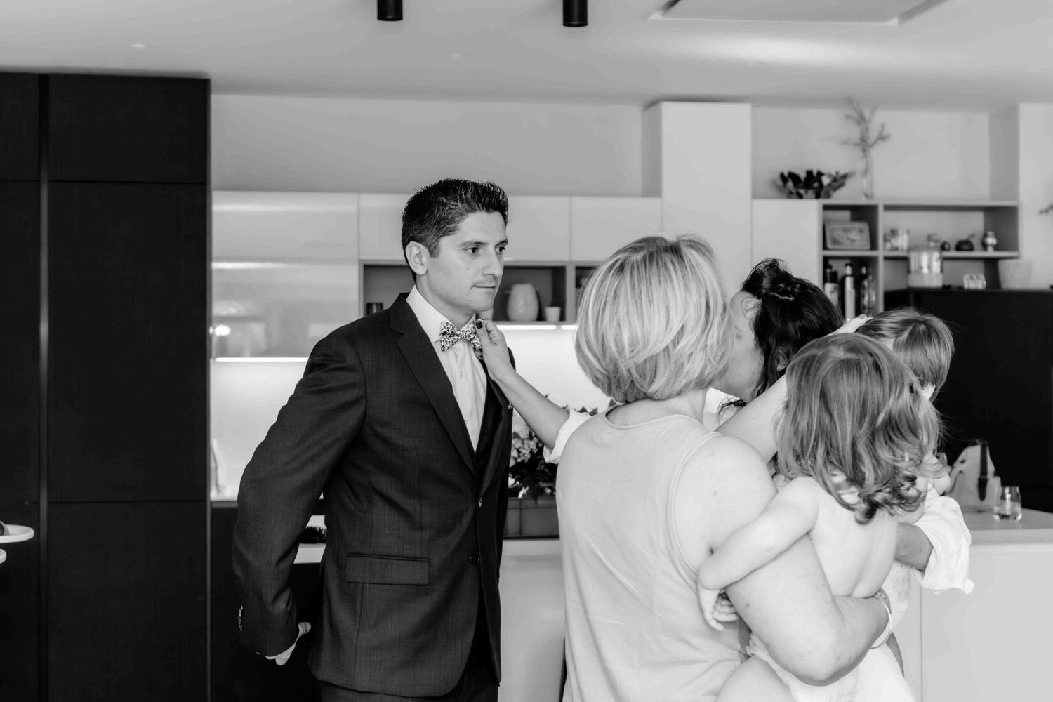 A black and white candid moment captured at a casual indoor gathering with people interacting in a kitchen setting.