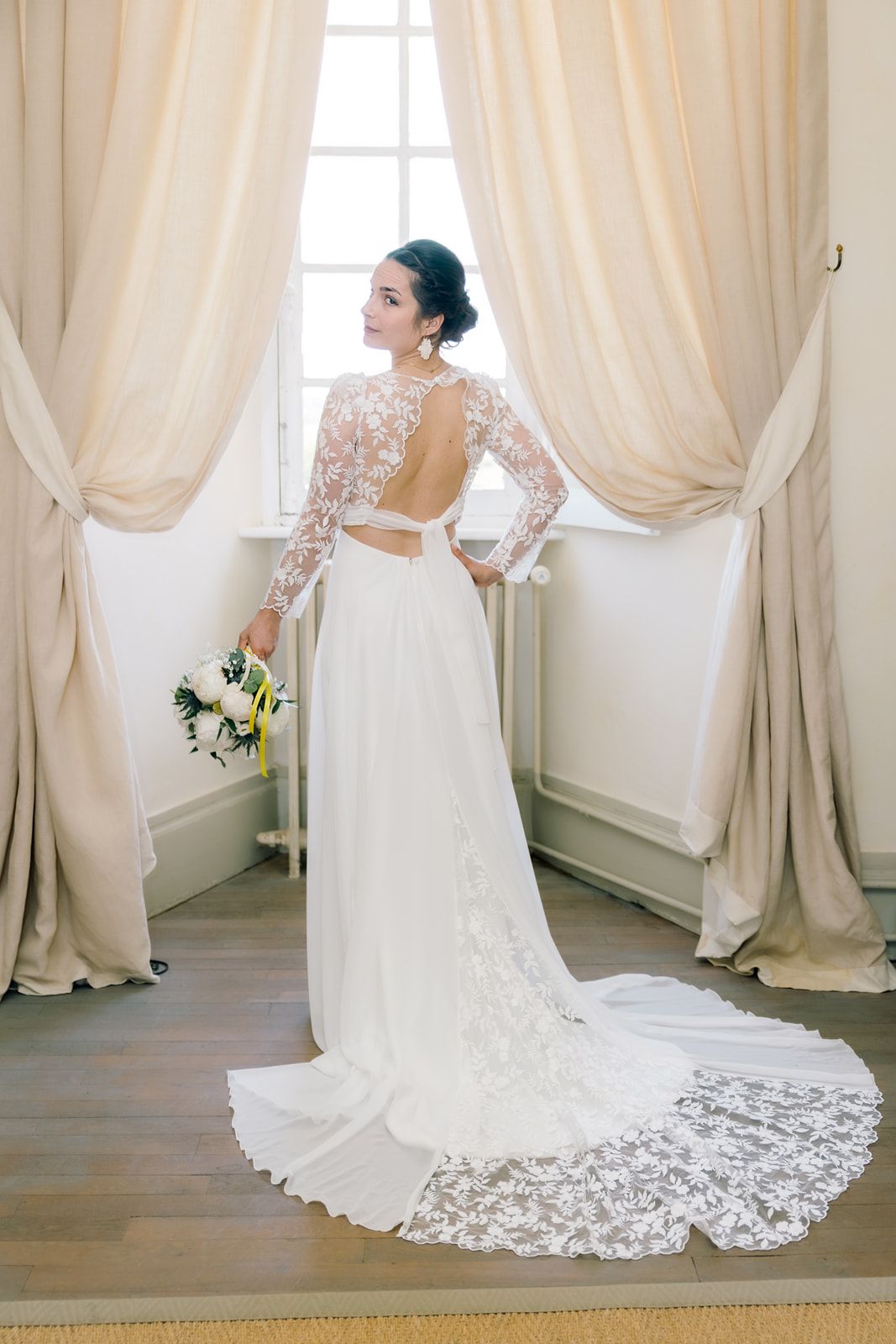 Bride in elegant backless lace wedding gown with train stands by draped window holding bouquet.