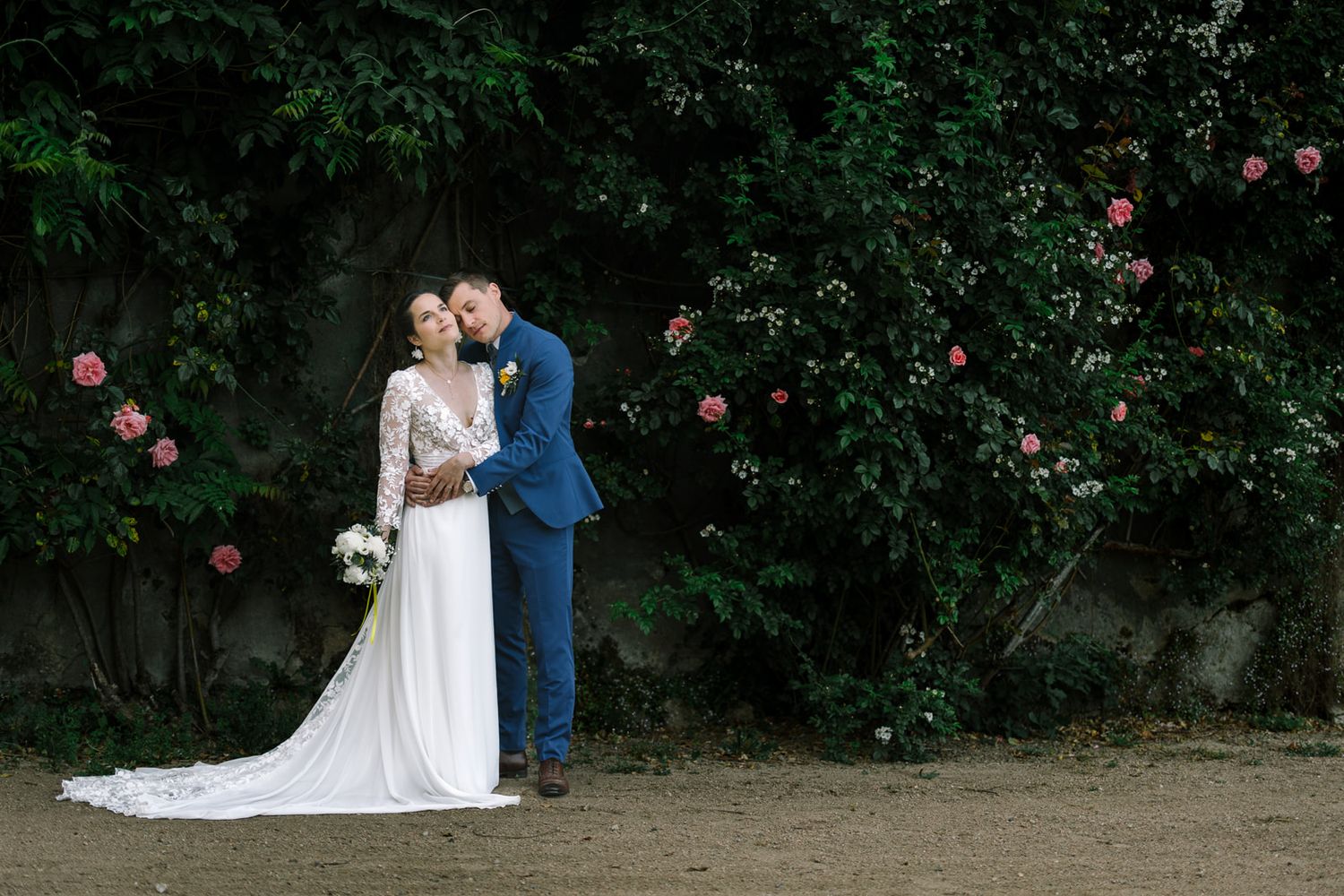 Bride and groom embrace before lush green wall with pink roses, bride in elegant white gown with train.
