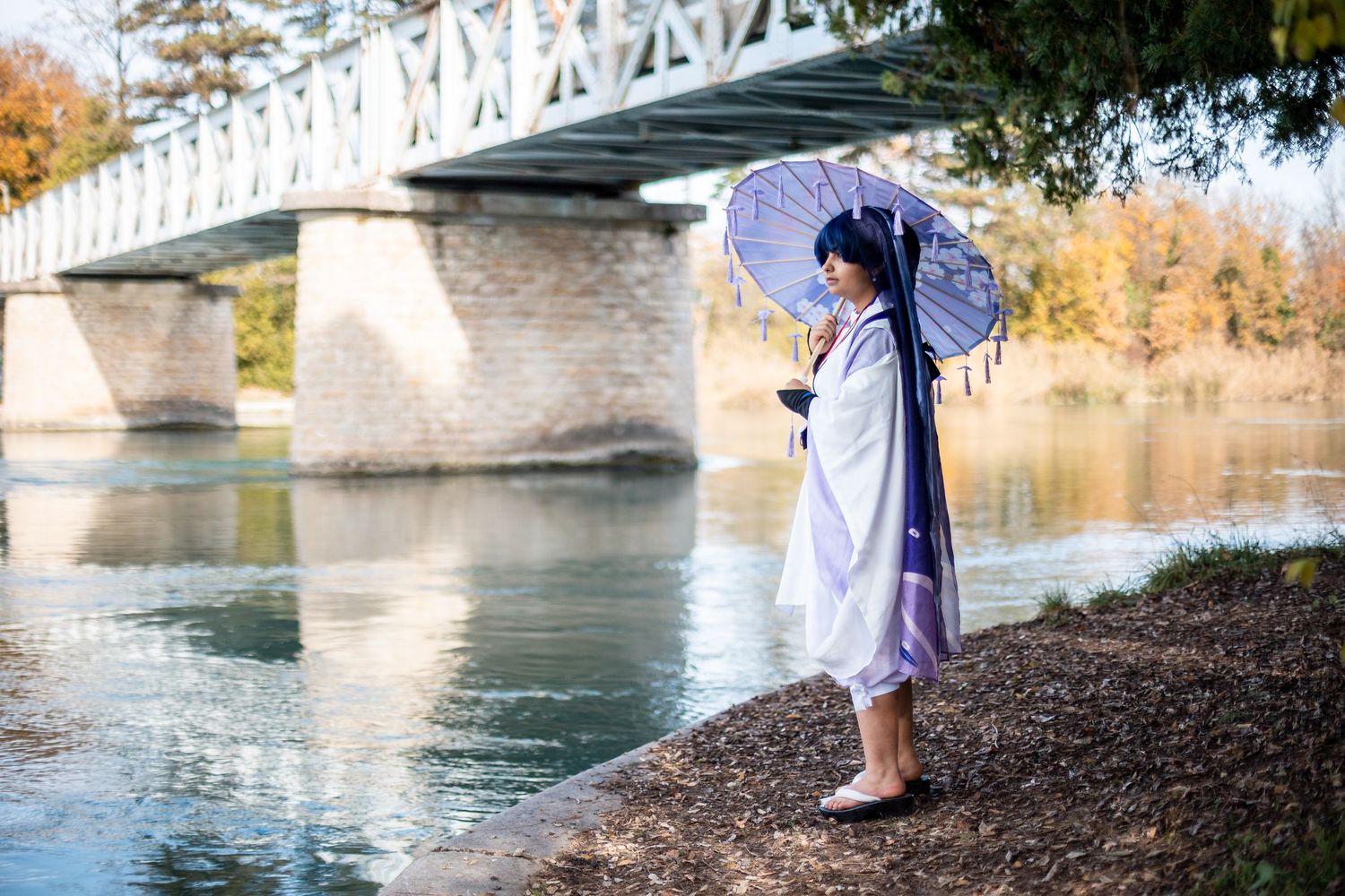 Figure in a lavender dress holds a lace parasol while standing near a white bridge over calm water.