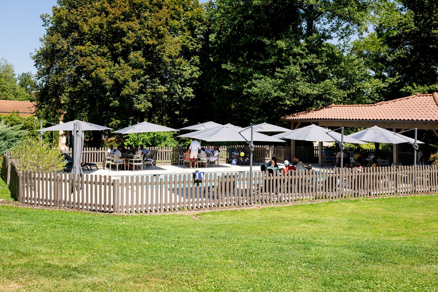 A series of white picket fences line a grassy outdoor area with umbrellas and seating visible in the background.