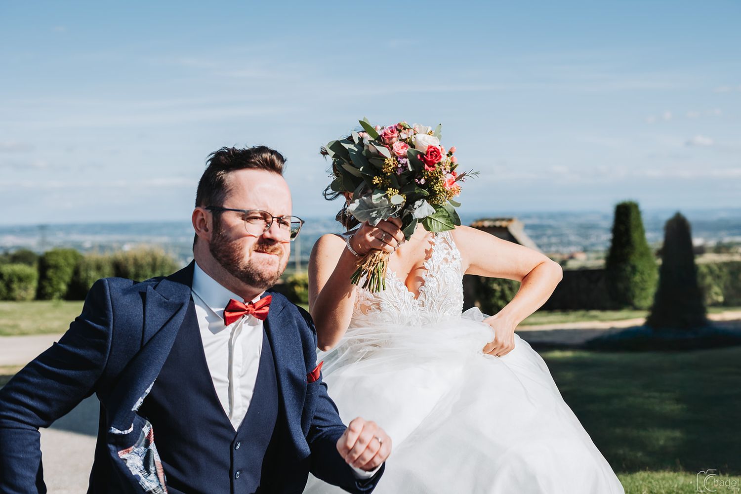A wedding couple poses outdoors on a sunny day with scenic mountain views in the background.