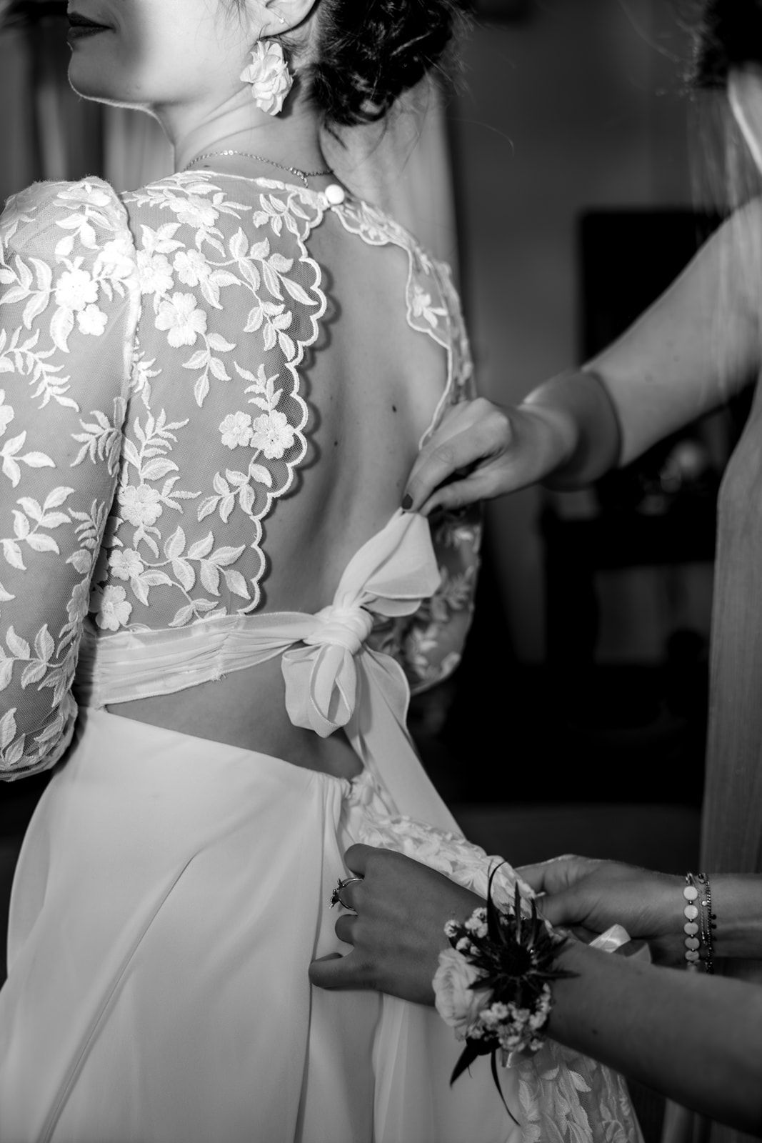 Black and white photo of a bride having her lace back wedding dress tied by bridesmaids.