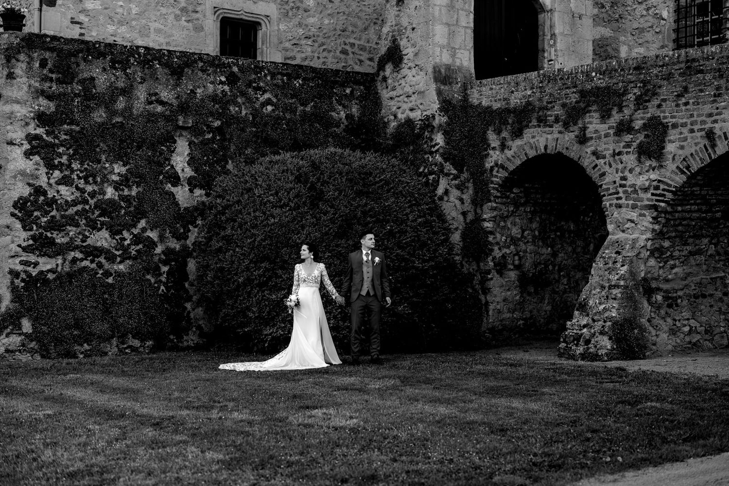 Black and white photo of bride and groom walking near stone arches and ivy-covered walls at a historic venue.