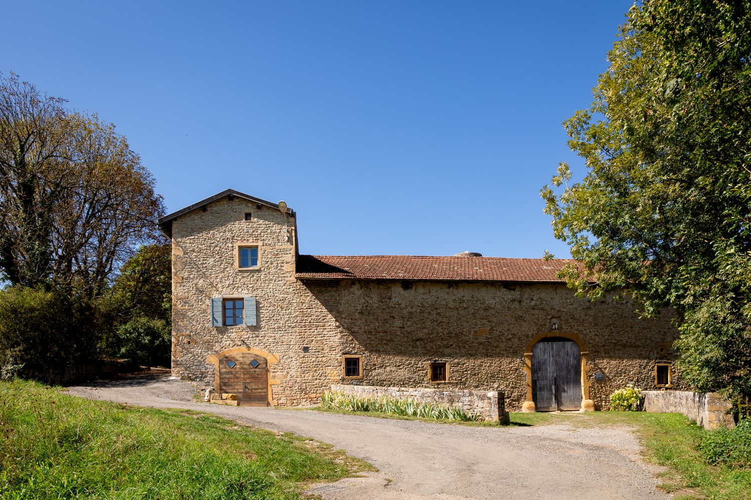Rustic stone farmhouse buildings with red tile roofs sit along a winding country path on a sunny day.