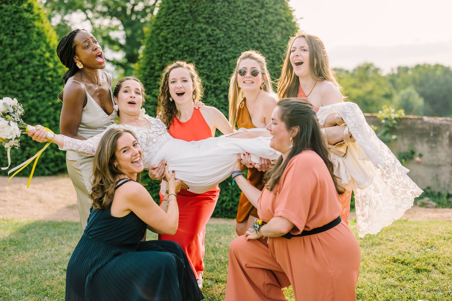 Bridesmaids joyfully lifting a bride in a white lace gown outdoors at a sunny garden wedding celebration.