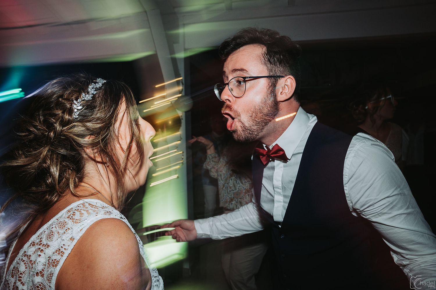 Two people dancing energetically at a wedding reception with colorful lights in the background.