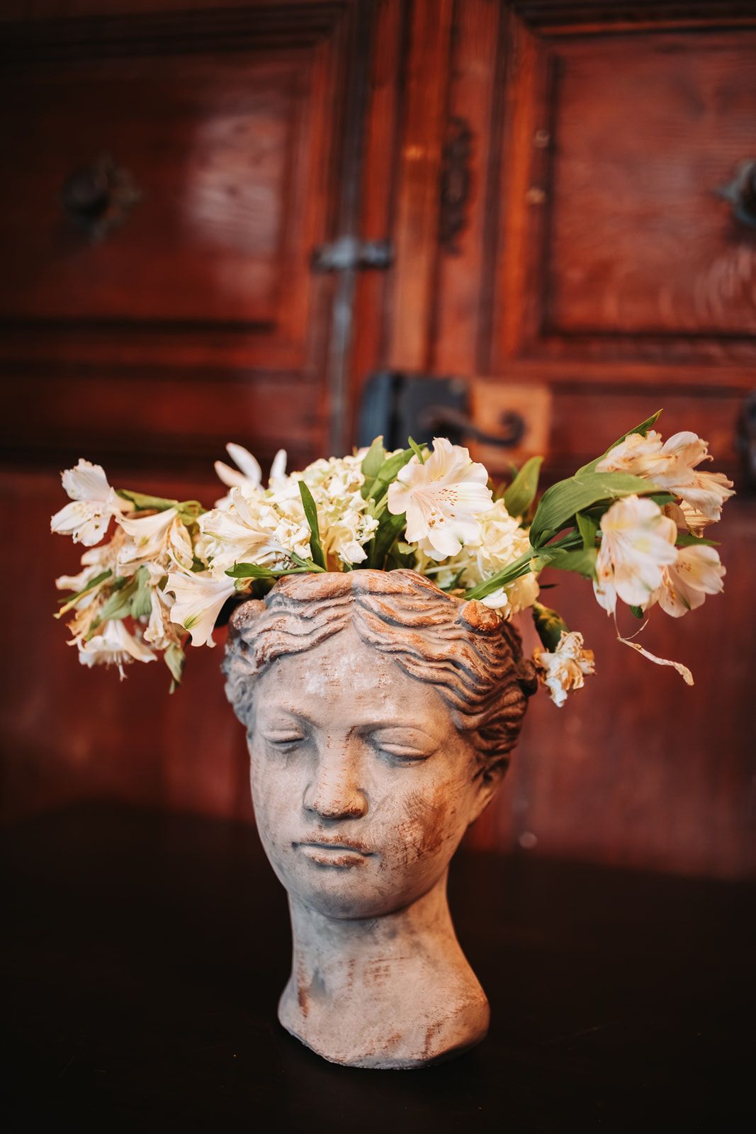 A classical stone bust vase with delicate white spring flowers arranged on top against a dark rustic background.