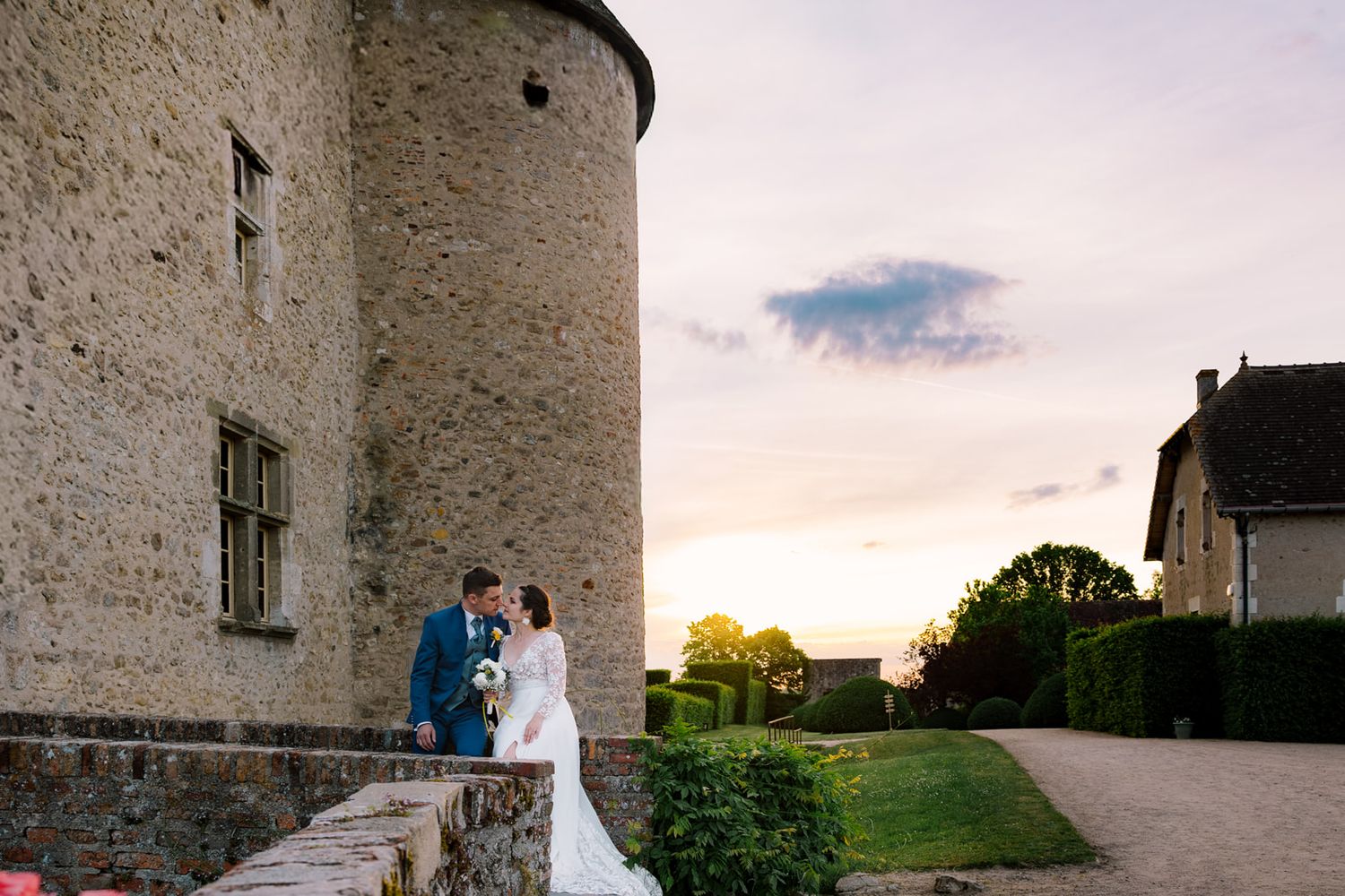 Bride and groom share a romantic kiss at sunset beside a historic stone castle tower at their wedding.