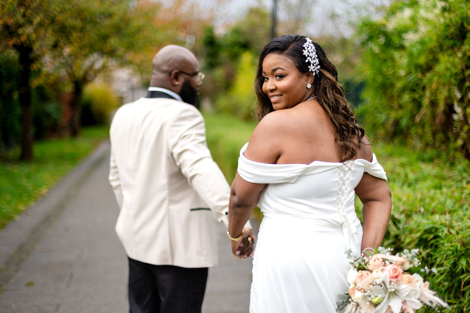 Black couple in wedding attire holding hands on a garden path, bride in off-shoulder gown looking back.