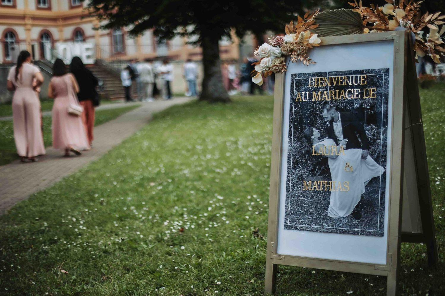 A wedding sign stands on a lawn with blurred figures in pink dresses walking in the background.