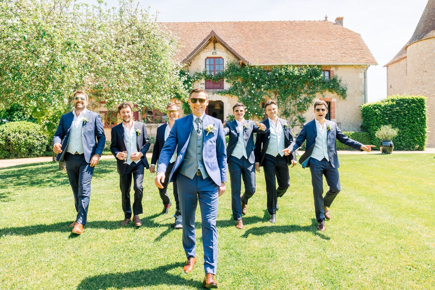 Groom leads groomsmen in matching blue suits across green lawn outside rustic French venue on sunny wedding day.